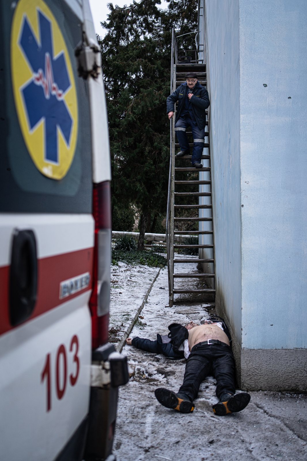 Una ambulancia junto a un hombre malherido tendido en el suelo, con otro hombre bajando una escalera de emergencia.